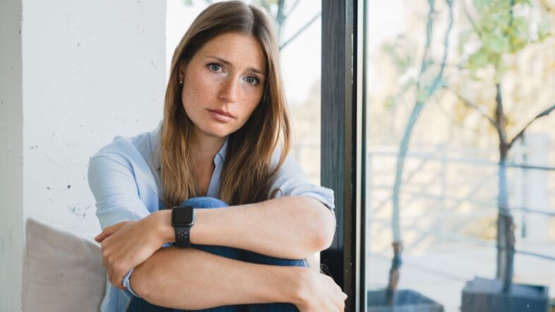 Depressed young woman thinking about breakup sitting by the window alone at home.