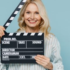 Elderly caucasian woman 50s wearing casual striped shirt looking camera holding classic black film making clapperboard isolated on plain pastel light blue color background.