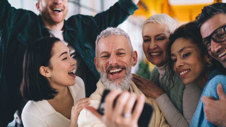 Cheerful male employee making positive selfie photos during day brainstorming with successful colleagues.