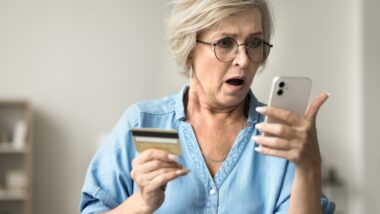 Shocked concerned mature woman in glasses holding credit card, staring at smartphone in bad surprise, getting stress, financial problems, money stealing.